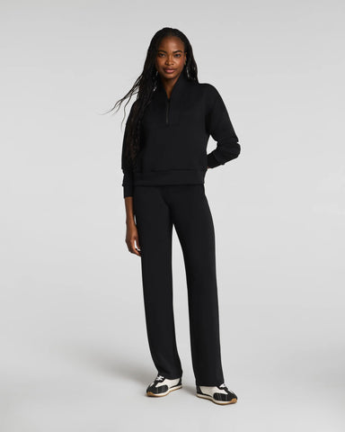 A woman modeling a black two-piece tracksuit consisting of a half-zip long-sleeve top and matching wide-leg pants, paired with black and white sneakers, against a light gray studio background.
