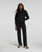 A woman modeling a black two-piece tracksuit consisting of a half-zip long-sleeve top and matching wide-leg pants, paired with black and white sneakers, against a light gray studio background.