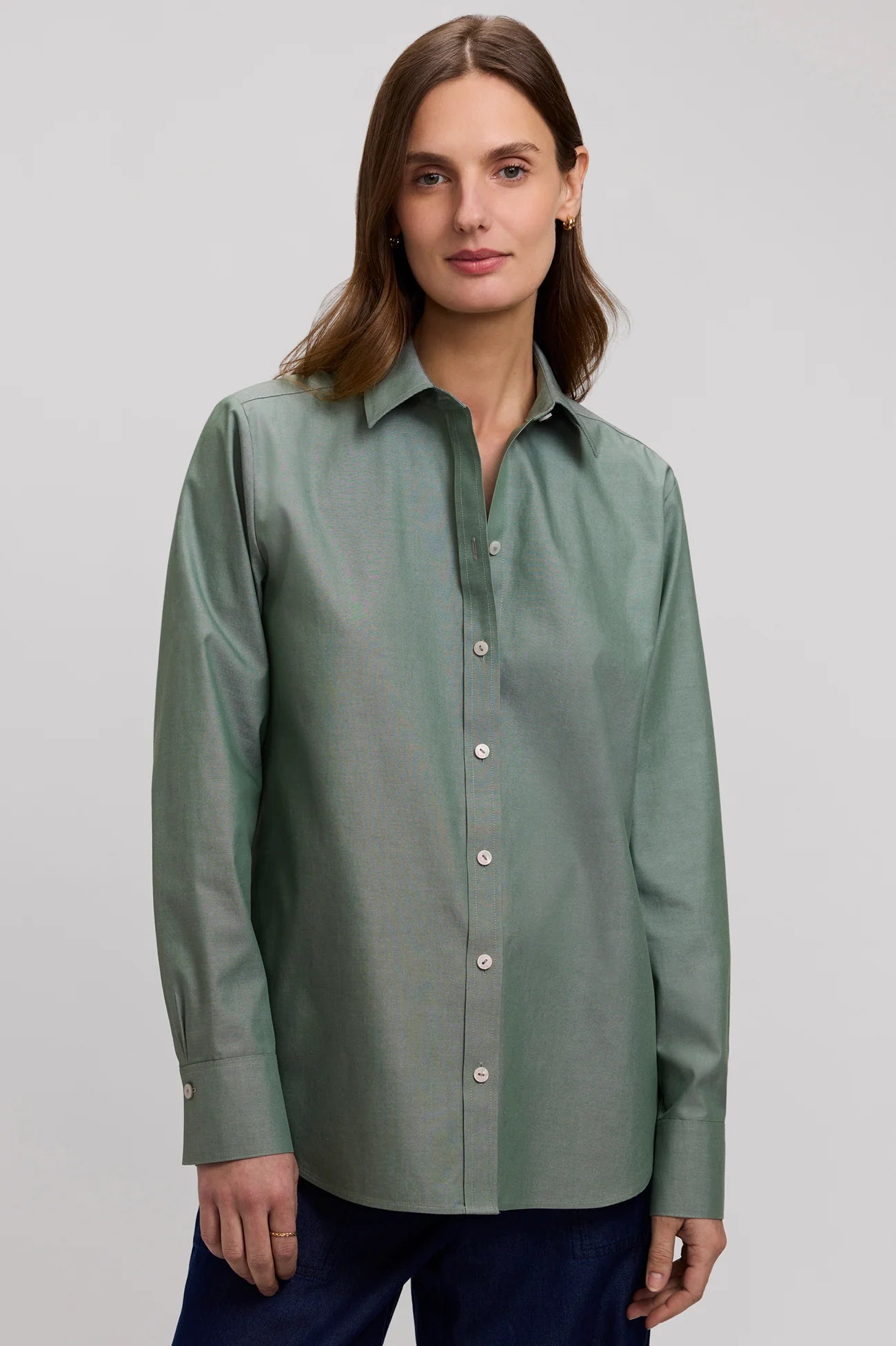 A woman wearing a sage green long-sleeve button-up shirt and navy trousers, standing against a neutral light gray studio background.
