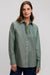 A woman wearing a sage green long-sleeve button-up shirt and navy trousers, standing against a neutral light gray studio background.