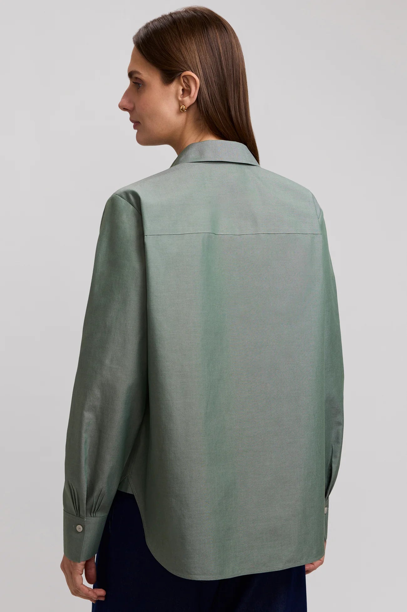 A woman wearing a sage green long-sleeve button-up shirt and dark blue pants, photographed against a light gray studio background.