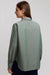 A woman wearing a sage green long-sleeve button-up shirt and dark blue pants, photographed against a light gray studio background.
