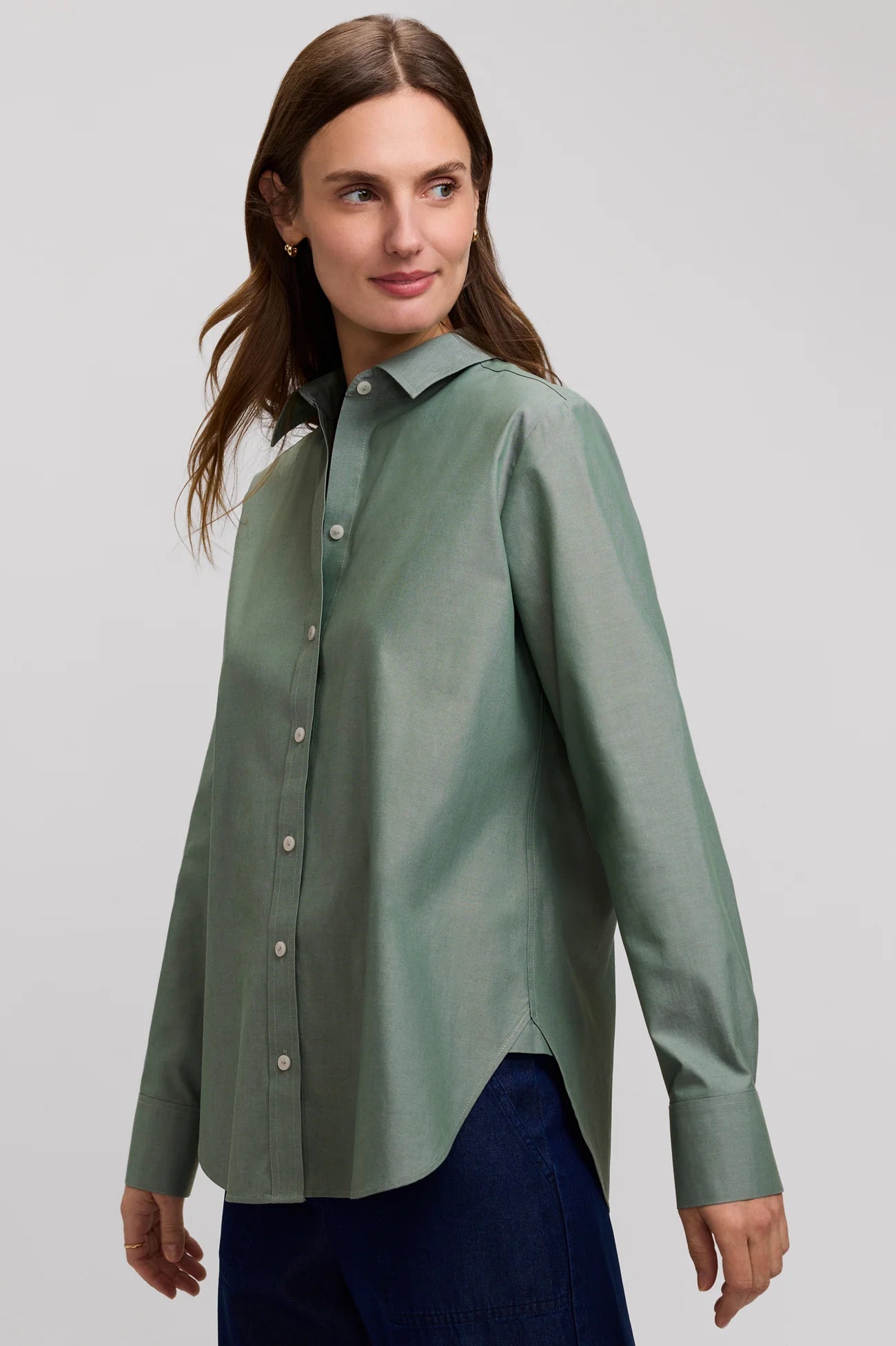 A woman wearing a sage green long-sleeve button-up shirt and dark blue pants, photographed against a light gray studio background.