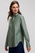 A woman wearing a sage green long-sleeve button-up shirt and dark blue pants, photographed against a light gray studio background.