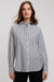 A woman wearing a long-sleeve button-up shirt with vertical navy and white stripes, paired with black pants, photographed against a neutral light gray studio background.