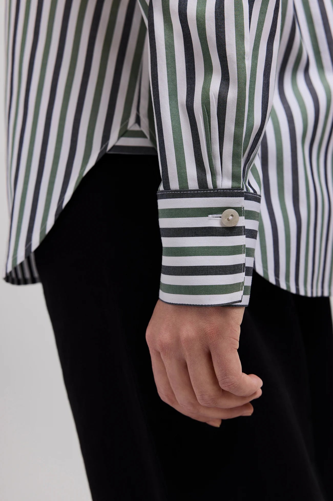 Close-up of a person wearing a striped button-up shirt with green, white, and black vertical stripes, paired with black trousers. The shirt sleeve is rolled up to show the cuff with a single button.
