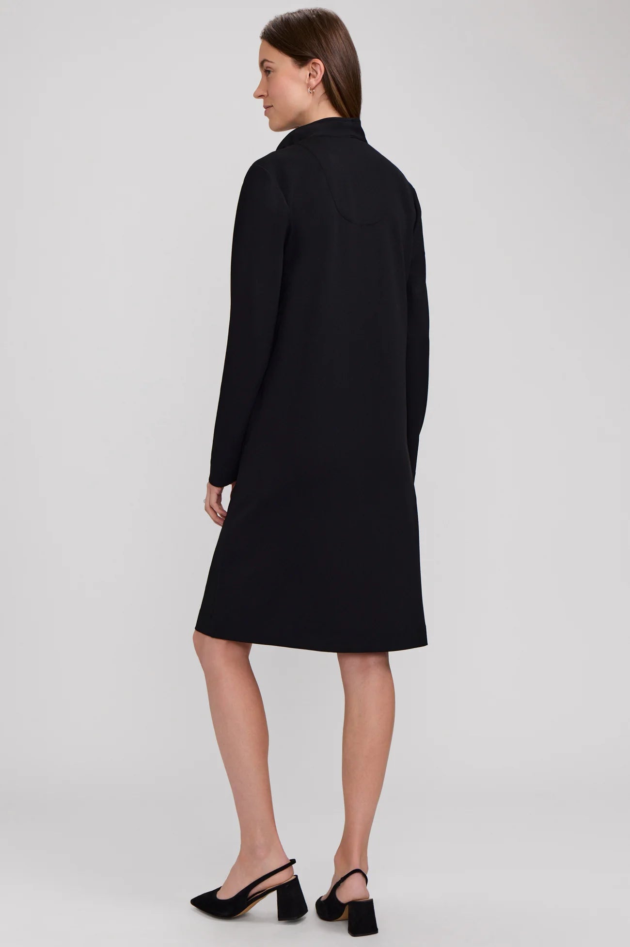 A woman modeling a black long-sleeve knee-length coat or dress from the back, paired with black slingback heels, against a light gray studio background.