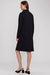 A woman modeling a black long-sleeve knee-length coat or dress from the back, paired with black slingback heels, against a light gray studio background.