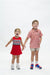 Two children modeling clothing against a plain white studio background. The child on the left wears a red sleeveless dress with 'GEORGIA' across the chest, and the child on the right wears a pink patterned short-sleeve polo shirt with beige shorts and colorful sneakers.