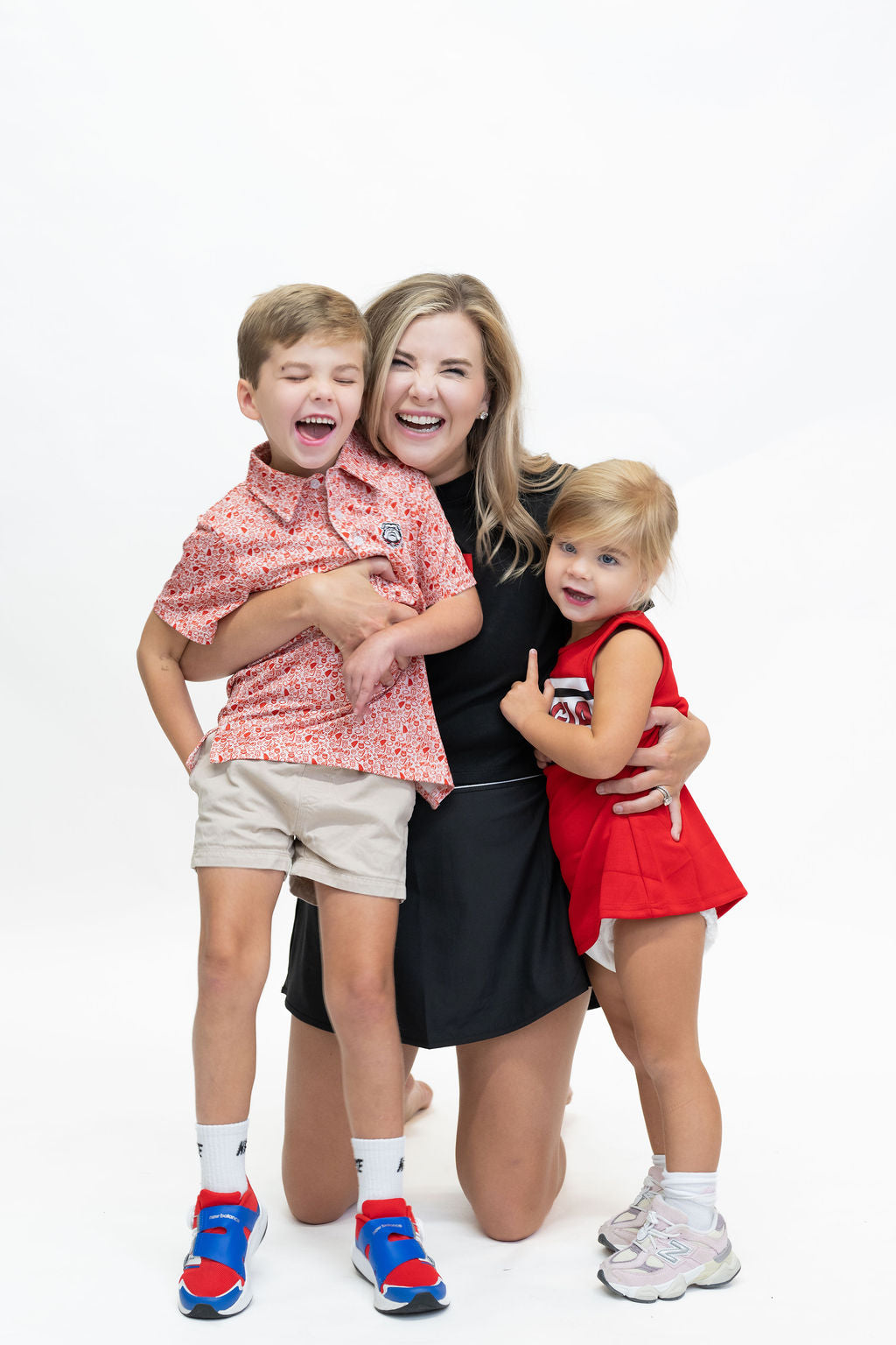 A woman and two children posing together against a solid white studio background. The woman is kneeling on one knee, wearing a black dress and smiling.