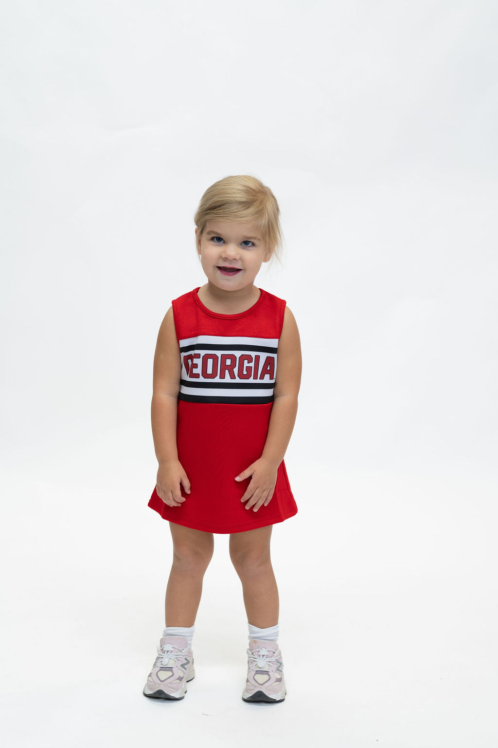 A young child stands against a plain white studio background wearing a red sleeveless dress with a horizontal stripe pattern across the chest featuring the word 'GEORGIA' in white with a red outline.