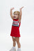 A toddler girl with blonde hair stands with her arms raised, wearing a red sleeveless athletic dress with 'GEORGIA' printed across the chest in white and black stripes, paired with white socks and sneakers, against a plain white studio background.