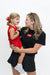 A woman in a black short-sleeve top and black skirt holding a young girl wearing a red sleeveless top and a red hair bow, against a plain white studio background. The woman's top has a red logo patch on the chest.
