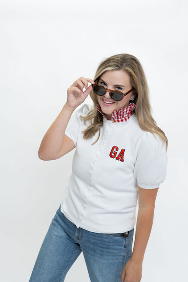 A woman modeling a white short-sleeve top with red 'GA' lettering on the chest, paired with blue jeans and a red-and-white gingham neck gaiter, against a plain white background.
