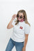 A woman modeling a white short-sleeve top with red 'GA' lettering on the chest, paired with blue jeans and a red-and-white gingham neck gaiter, against a plain white background.