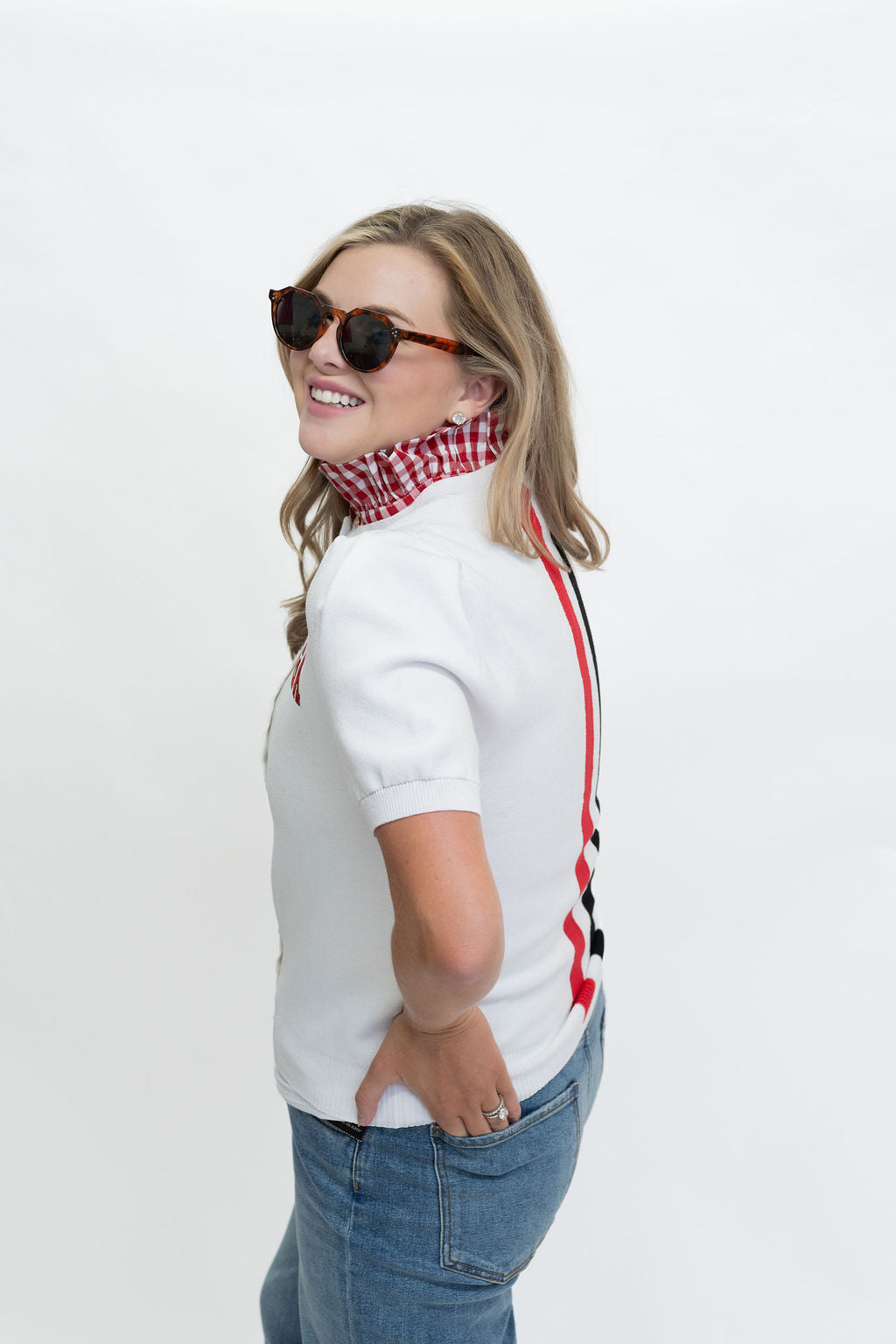 A woman wearing round sunglasses, a red and white checkered neck gaiter, a white short-sleeve top with red and black side stripes, and blue jeans, photographed against a plain white studio background.