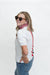A woman wearing round sunglasses, a red and white checkered neck gaiter, a white short-sleeve top with red and black side stripes, and blue jeans, photographed against a plain white studio background.
