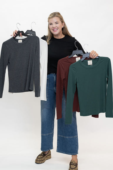 A woman stands against a white studio background holding up four long-sleeve knit tops on hangers, each in a different color: dark gray, white, burgundy, and forest green. She is wearing a black top, wide-leg blue jeans, and brown loafers.