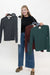 A woman stands against a white studio background holding up four long-sleeve knit tops on hangers, each in a different color: dark gray, white, burgundy, and forest green. She is wearing a black top, wide-leg blue jeans, and brown loafers.