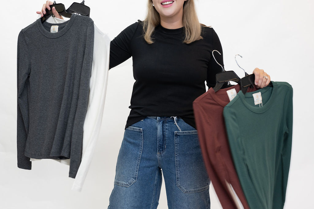 A woman stands against a white studio background holding up three long-sleeve crewneck knit tops on hangers. The tops are in dark gray, white, burgundy, and forest green.