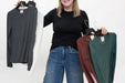 A woman stands against a white studio background holding up three long-sleeve crewneck knit tops on hangers. The tops are in dark gray, white, burgundy, and forest green.