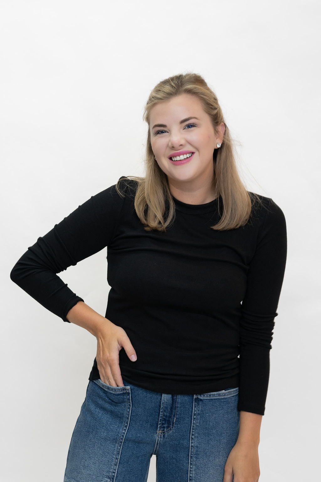 A smiling blonde woman wearing a fitted black long-sleeve top and blue jeans, standing against a plain white studio background.
