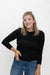 A smiling blonde woman wearing a fitted black long-sleeve top and blue jeans, standing against a plain white studio background.