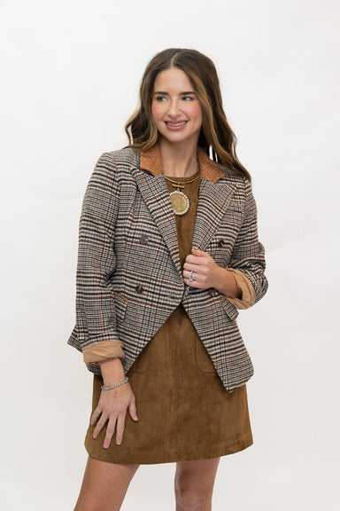 A woman modeling a brown and beige plaid blazer with tan suede-like trim over a brown dress, standing against a plain white studio background.