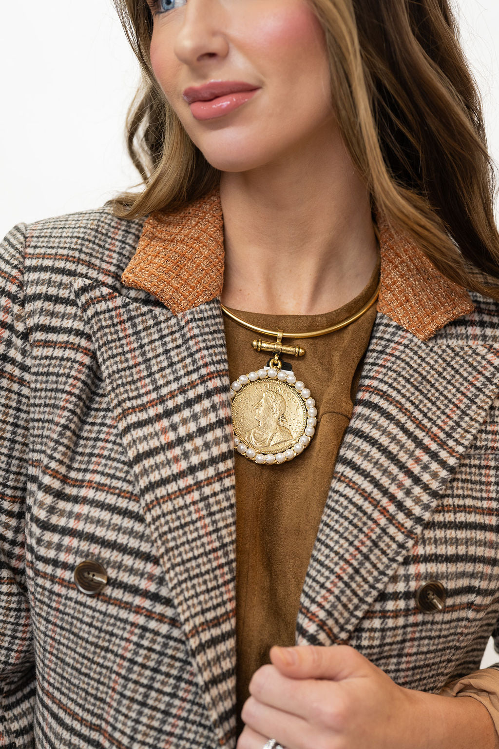 A close-up of a woman wearing a brown and beige plaid blazer with a tan collar, a brown top, and a gold necklace featuring a large coin pendant.