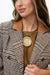 A close-up of a woman wearing a brown and beige plaid blazer with a tan collar, a brown top, and a gold necklace featuring a large coin pendant.