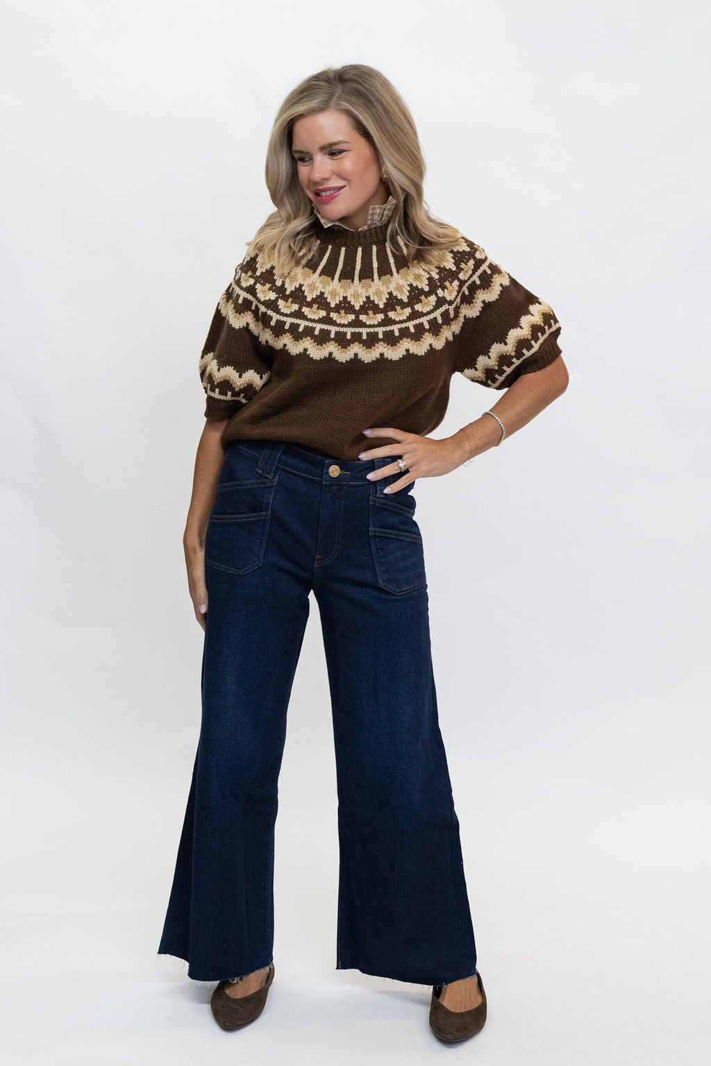 A woman modeling a brown short-sleeve knit top with a beige geometric pattern around the yoke, paired with dark blue wide-leg jeans and brown shoes, against a plain white studio background.