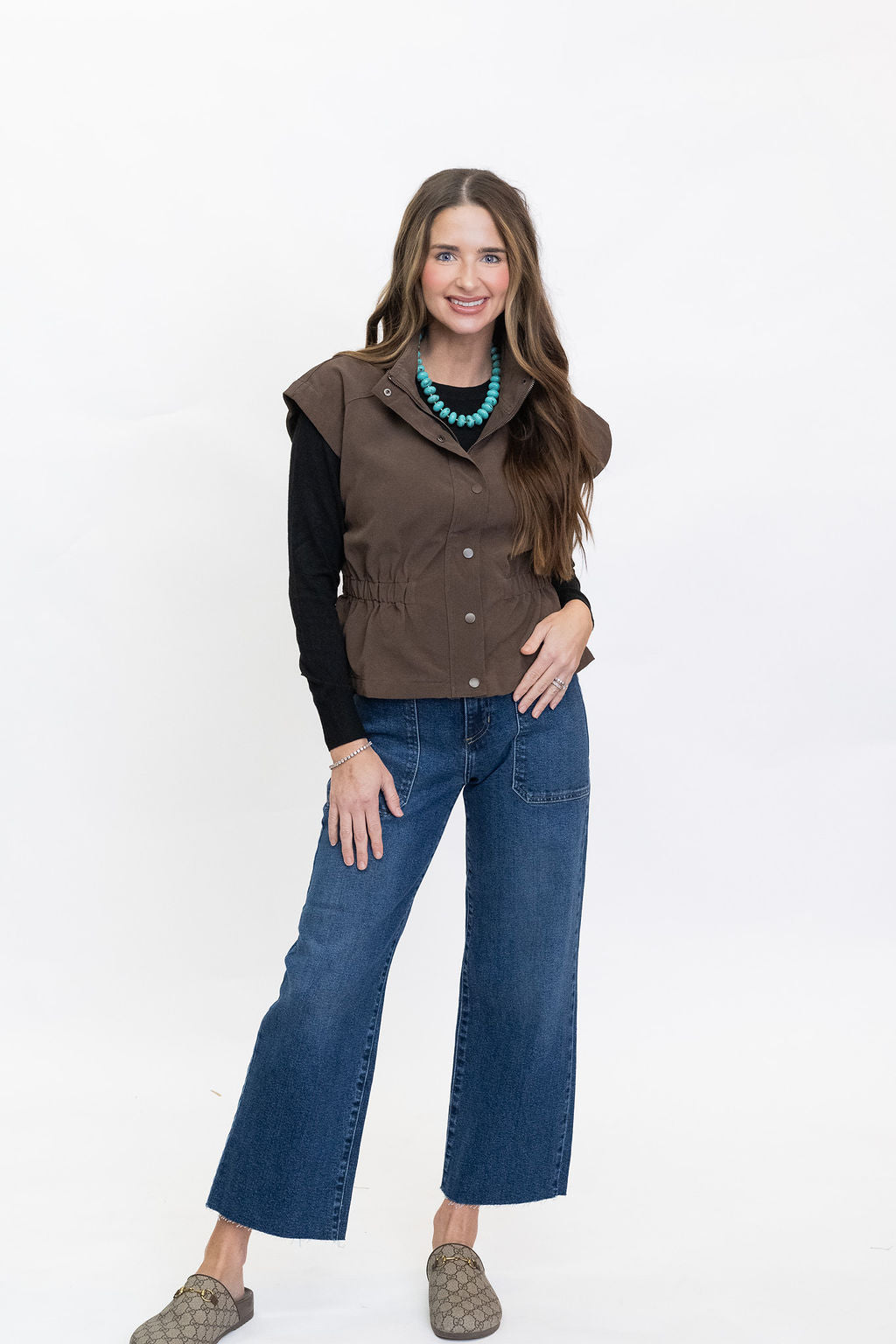 A woman modeling a sleeveless brown button-front vest over a black long-sleeve top, paired with blue jeans and brown slip-on shoes, against a plain white studio background.
