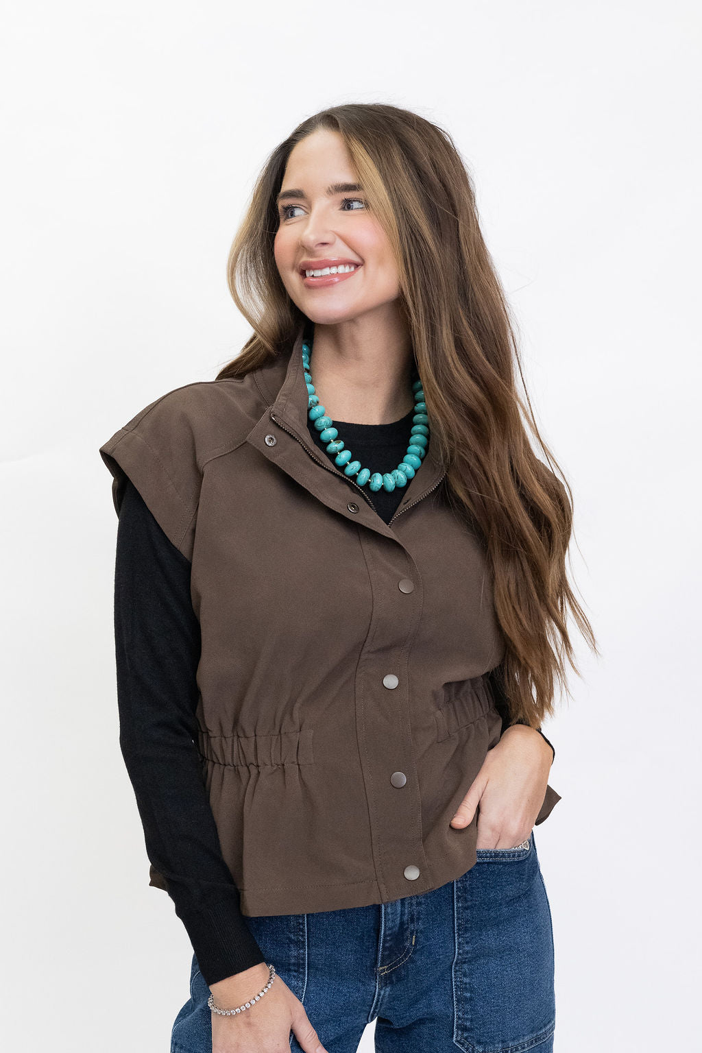 A woman modeling a sleeveless brown button-front vest over a black long-sleeve top and blue jeans, against a plain white studio background.