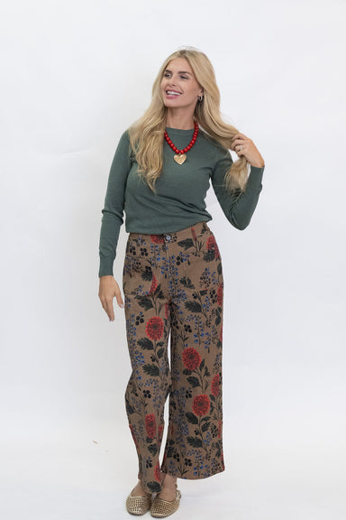 A woman modeling a two-piece outfit consisting of a sage green long-sleeve top and brown wide-leg trousers with a red and blue floral pattern, paired with a red beaded necklace and woven espadrilles, against a white studio background.