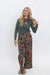 A woman modeling a two-piece outfit consisting of a sage green long-sleeve top and brown wide-leg trousers with a red and blue floral pattern, paired with a red beaded necklace and woven espadrilles, against a white studio background.