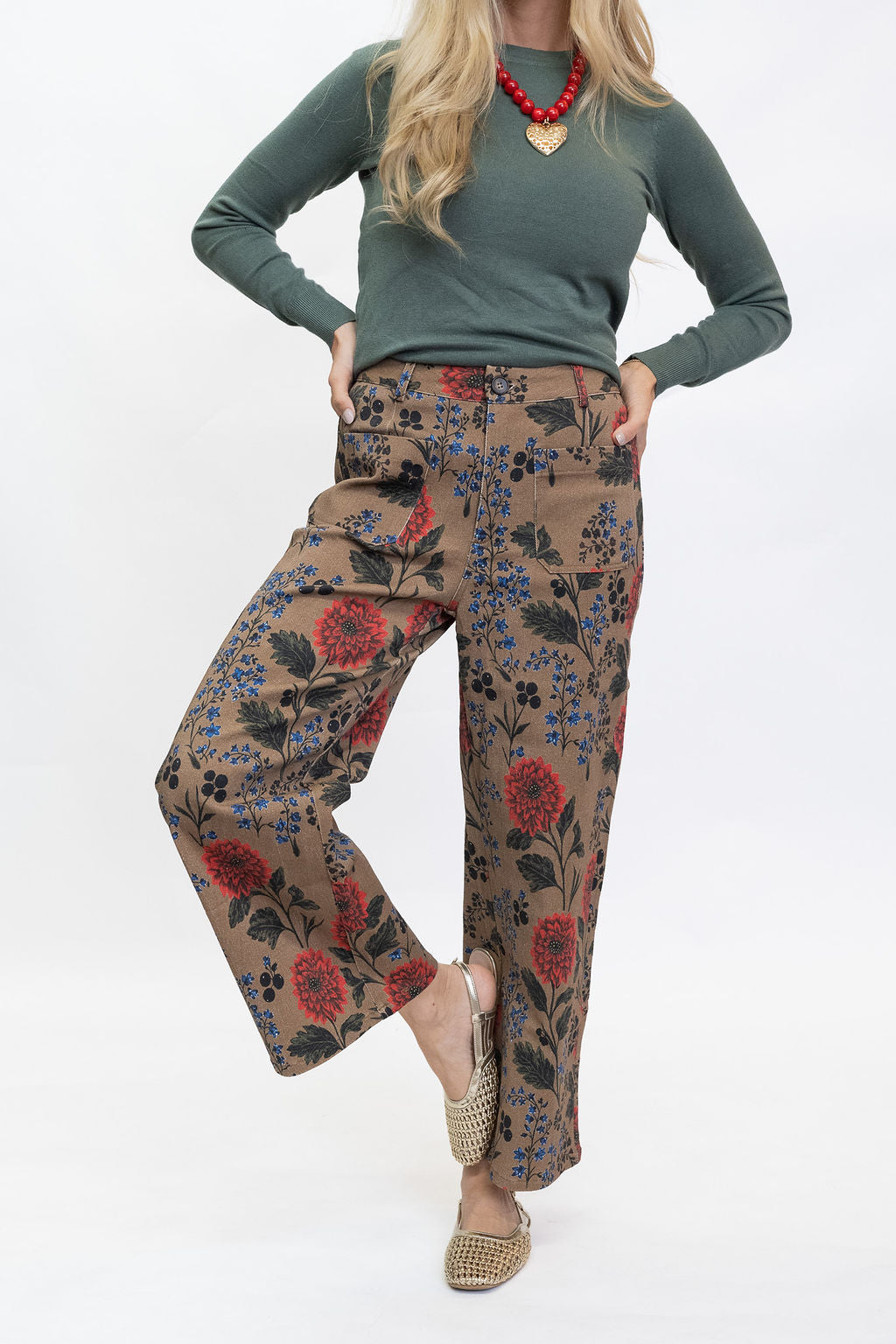 A model wearing brown wide-leg pants with a red, blue, and black floral pattern, styled with a sage green long-sleeve top and beige woven flat shoes against a white studio background.