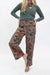 A model wearing brown wide-leg pants with a red, blue, and black floral pattern, styled with a sage green long-sleeve top and beige woven flat shoes against a white studio background.