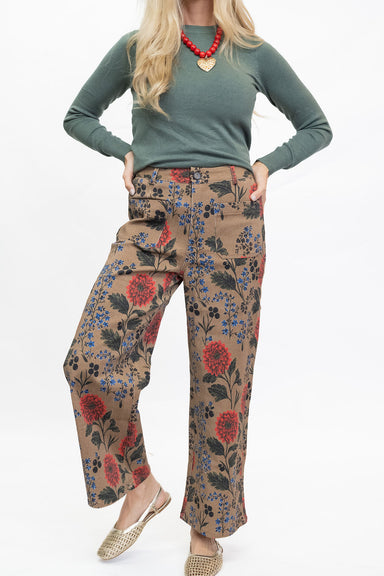 A model wearing a sage green long-sleeve top and wide-leg brown pants with a red and blue floral print, paired with a red beaded necklace and woven flat shoes, against a plain white background.