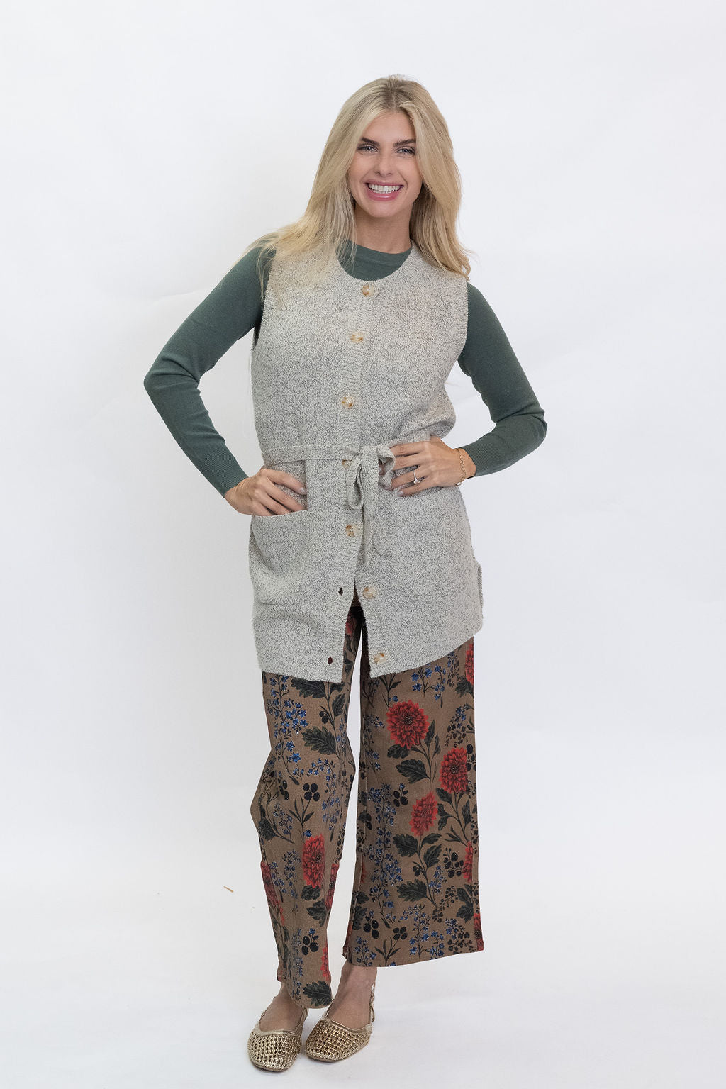 A woman modeling a light gray sleeveless button-front vest over a dark green long-sleeve top and brown wide-leg pants with a red and blue floral pattern, standing against a plain white studio background.