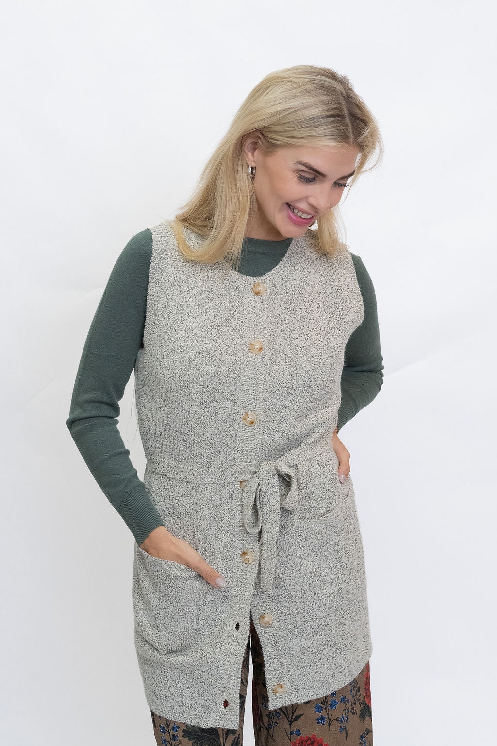 A woman modeling a light gray heathered knit sleeveless vest with a tie waist and front buttons, worn over a dark green long-sleeve shirt against a plain white studio background.