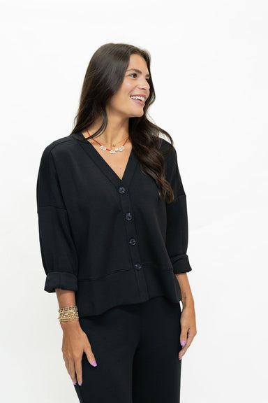 A woman wearing a black button-front cardigan top with three-quarter sleeves and black pants, photographed against a plain white studio background.