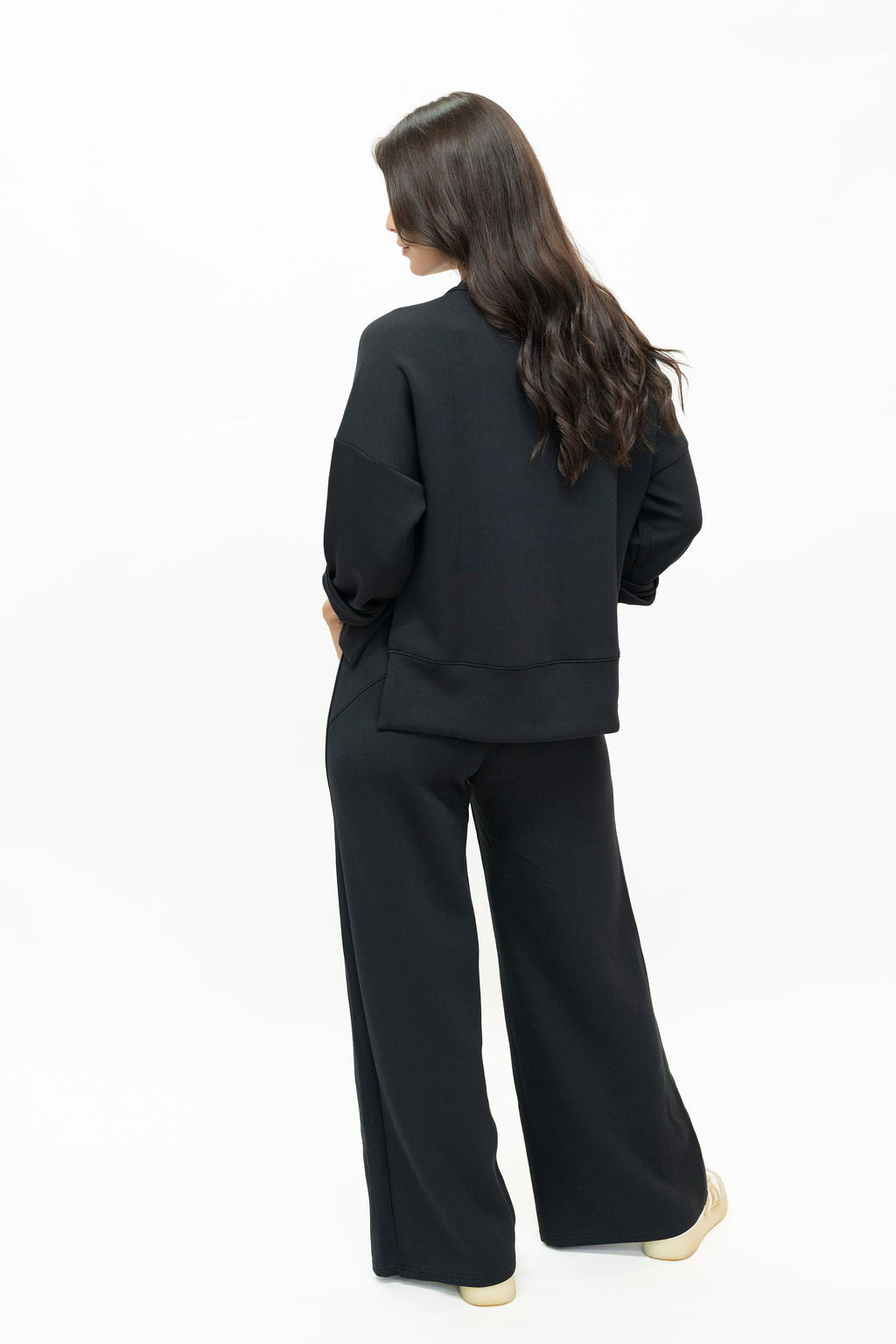 A woman with long dark hair is shown from the back wearing a black cropped long-sleeve top and matching wide-leg trousers, standing against a plain white studio background.