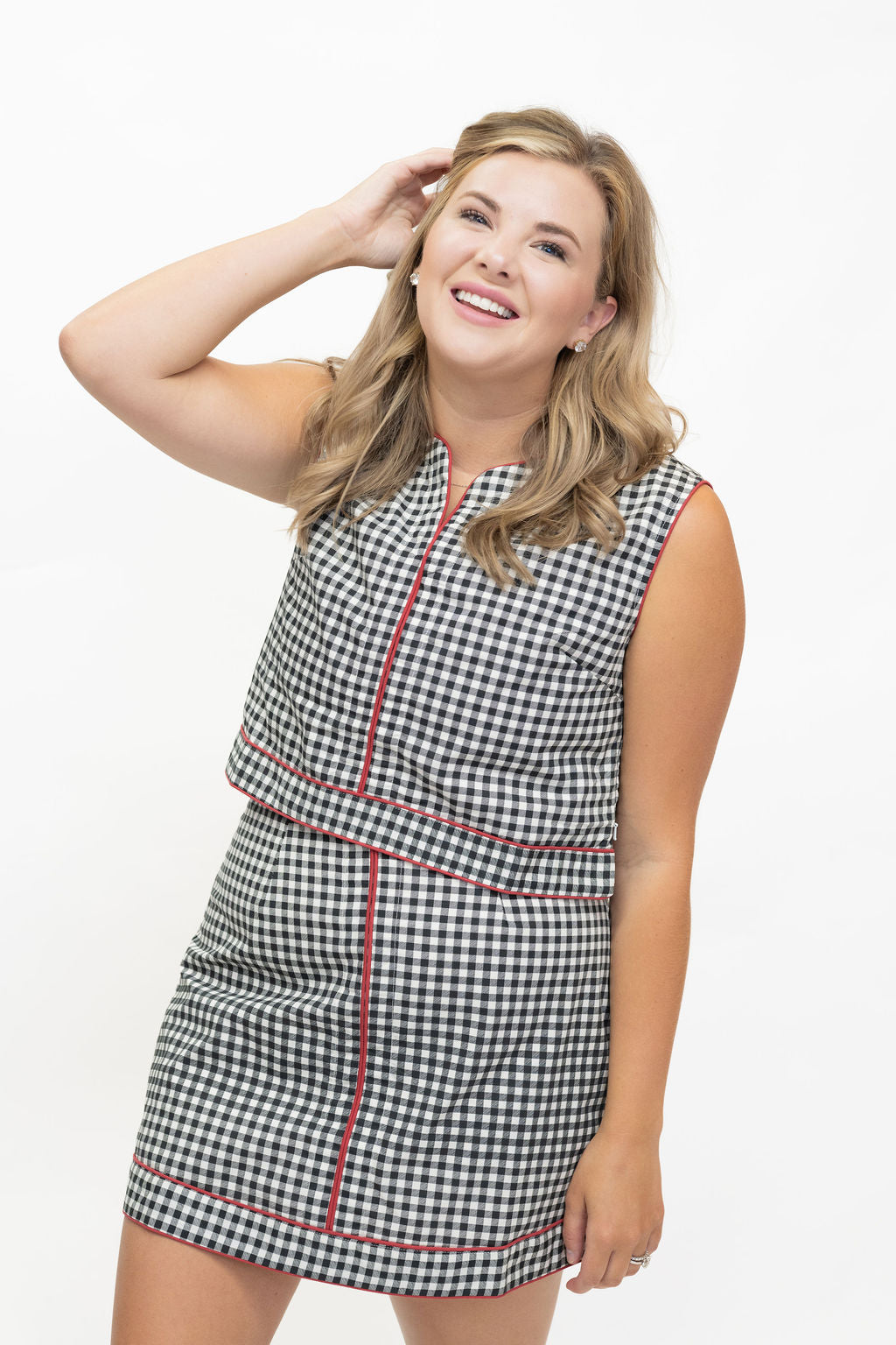 A woman modeling a sleeveless black and white gingham mini dress with red piping details, posing against a solid white studio background.