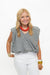 A smiling blonde woman wearing a black and white gingham sleeveless top, white pants, a chunky red beaded necklace, and gold bracelets, standing against a plain white background.