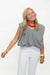 A smiling blonde woman wearing a black and white gingham sleeveless top, white pants, a chunky red beaded necklace, and gold bracelets, standing against a plain white studio background.