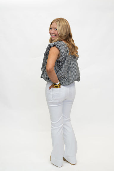 A woman with blonde hair is shown from a back-three-quarter angle, wearing a black and white gingham sleeveless top, white flared pants, and beige shoes, standing against a plain white studio background.