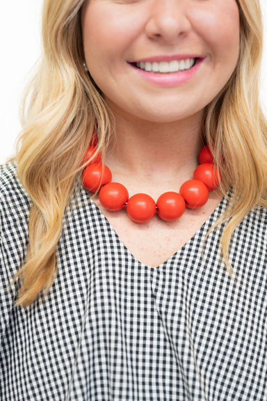 A close-up of a smiling woman wearing a chunky orange-red beaded necklace and a black-and-white gingham top against a plain white background.