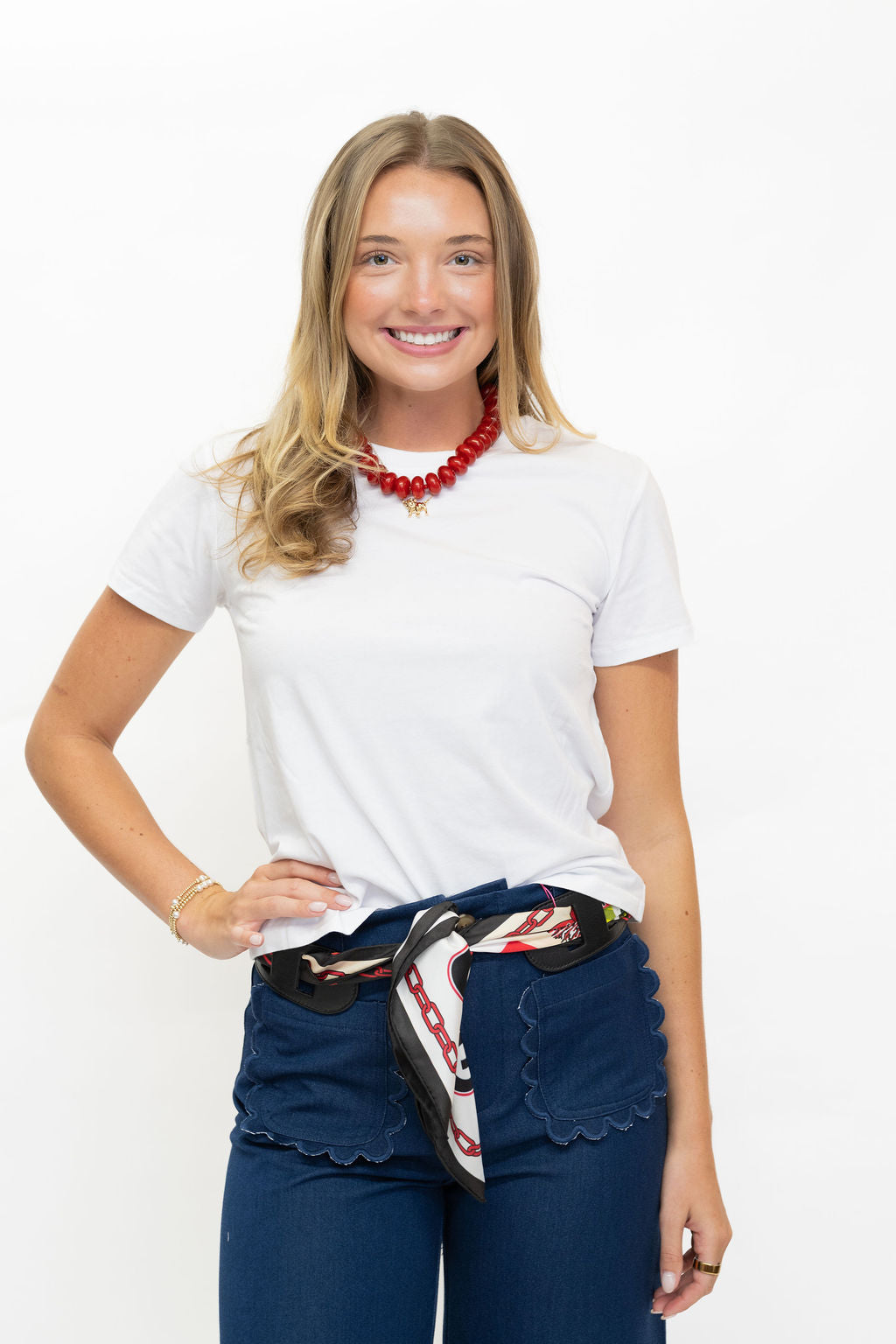 A smiling blonde woman wearing a white crewneck t-shirt, dark blue jeans with a patterned scarf tied at the waist, and a red beaded necklace, standing against a plain white background.