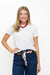 A smiling blonde woman wearing a white crewneck t-shirt, dark blue jeans with a patterned scarf tied at the waist, and a red beaded necklace, standing against a plain white background.