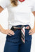 A close-up of a person wearing a white T-shirt, dark blue jeans with scalloped front pockets, and a patterned black, white, red, and gray scarf tied around the waist like a belt. The person is also wearing a red beaded necklace and gold bracelets against a plain white background.